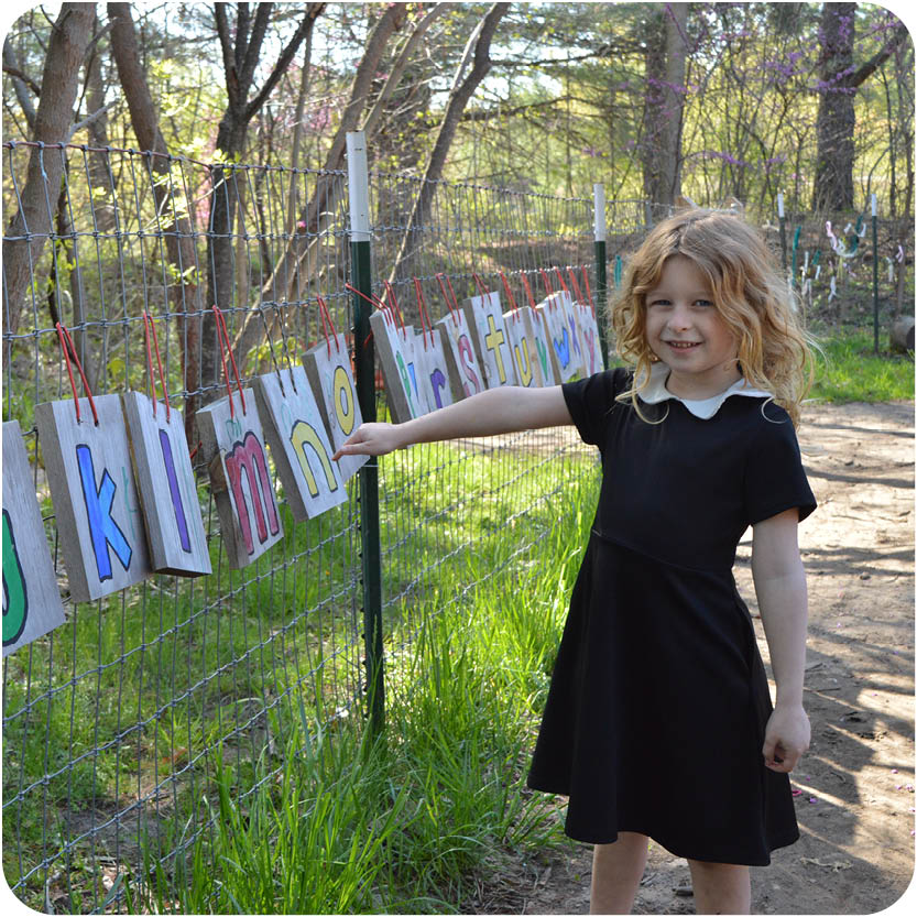 Story Hour - young girl in black dress pointing to hand made alphabet hanging on strings outside