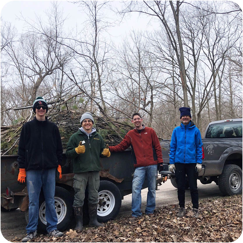 Weekend Land Maintenance volunteers pose smiling in front of a trailer full of sticks