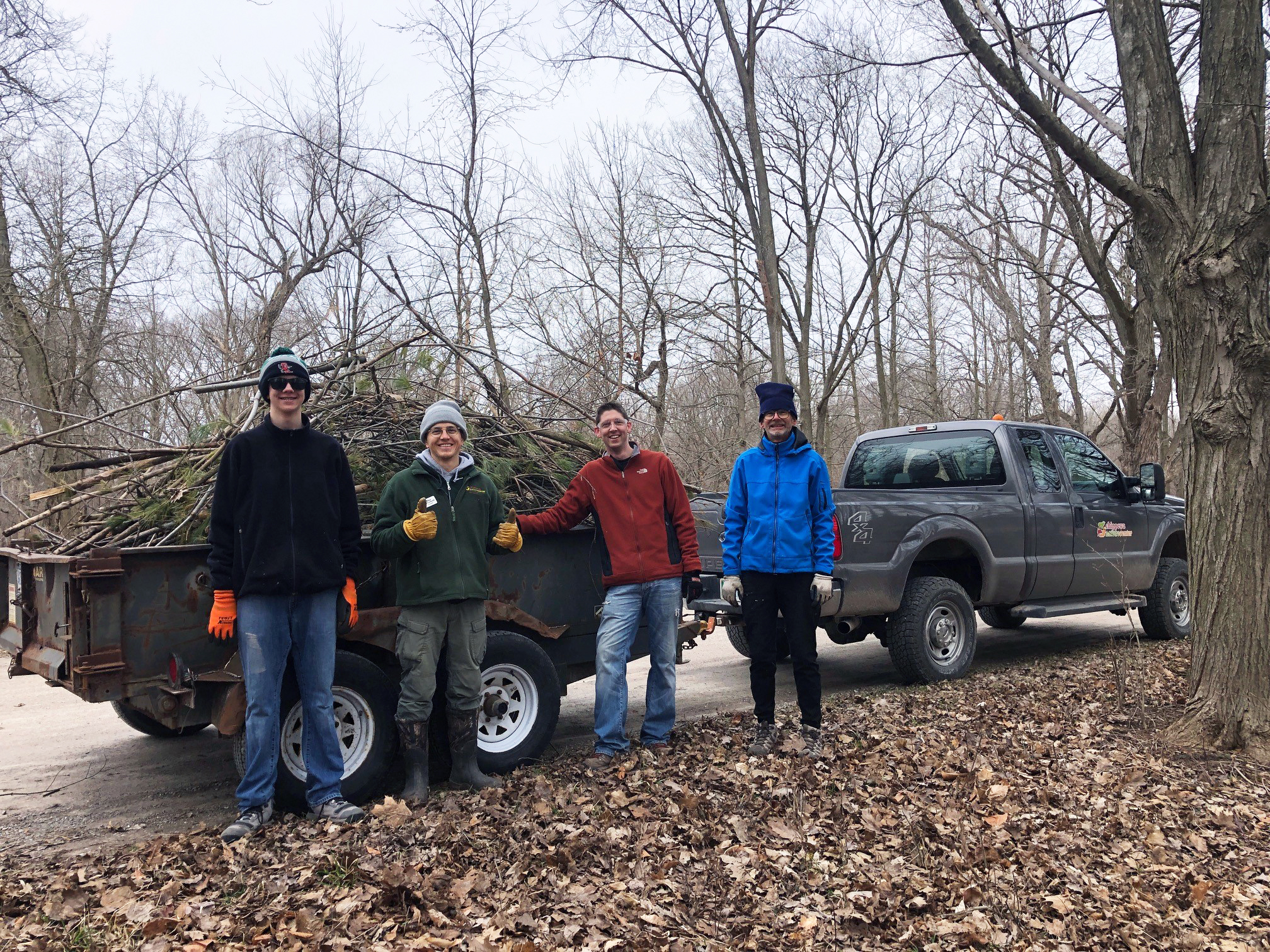 Weekend Land Maintenance volunteers pose smiling in front of a trailer full of sticks