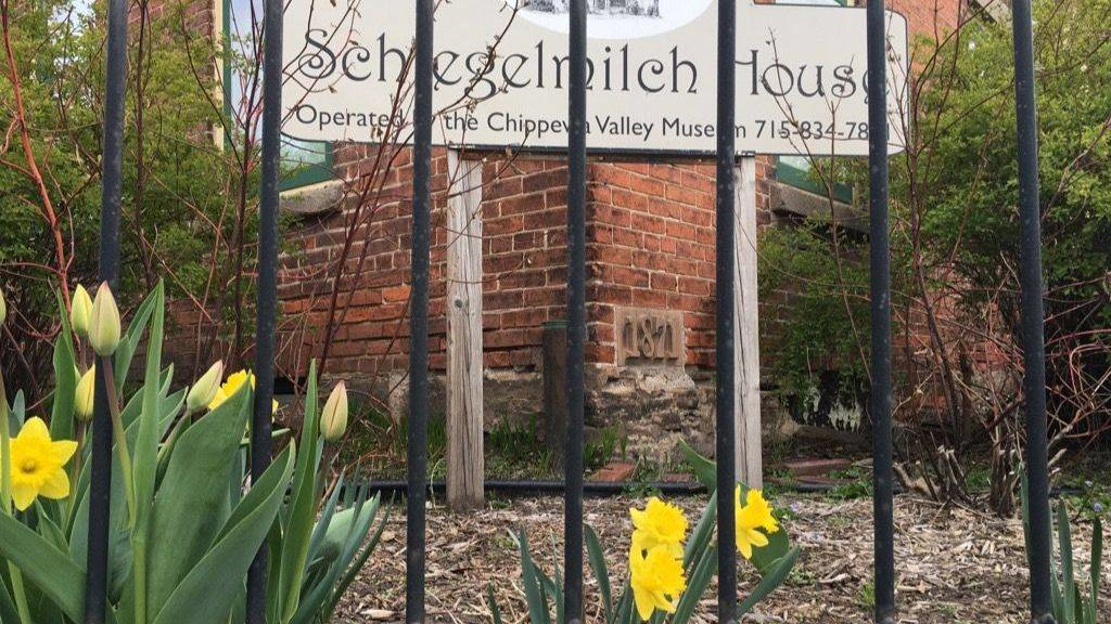 ground-level view through wrought iron fencing of a sign saying "Schlegelmilch House operated by the Chippewa Valley Museum 715-834-7871." Sign stands in front of the corner of a brick building with windows visible to each side. Yellow daffodils bloom in the foreground.