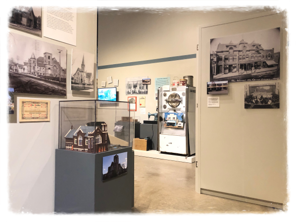 view of museum exhibit with model scale church in display case in foreground. Black and white images on three walls. Old fashioned refrigerator and sewing machine viewable in background.